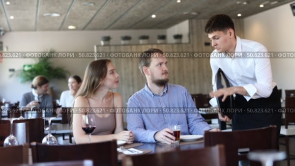 Couple resting, sitting in restaurant and ordering food. Waiter guy tells young women visitors of cafe about dish from chef. Стоковое видео, видеограф Яков Филимонов / Фотобанк Лори
