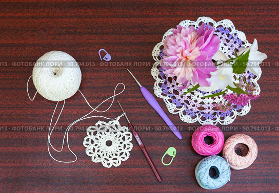 Crocheting lace napkin. Bouquet of flowers, crochet hooks, skeins of colored cotton yarn, pins on wooden table. DIY concept. Crochet as a hobby. Copy space, flat lay, close-up, top view. Стоковое фото, фотограф Виктория Катьянова / Фотобанк Лори