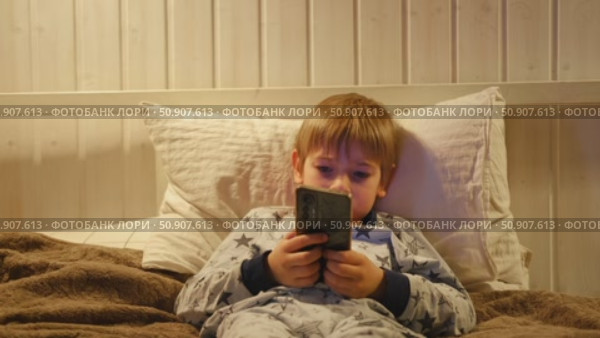 Cute kid boy in pajamas lying in bed watching smartphone, smiling. Children's screen time. Child plays games using phone at the evening or night. Social media addiction. Childhood, modern technology. Стоковое видео, видеограф Марина Демидюк / Фотобанк Лори