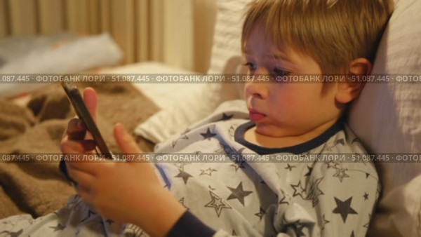 Cute kid boy in pajamas lying in bed watching smartphone, smiling. Children's screen time. Child plays games using phone at the evening or night. Social media addiction. Childhood, modern technology. Стоковое видео, видеограф Марина Демидюк / Фотобанк Лори