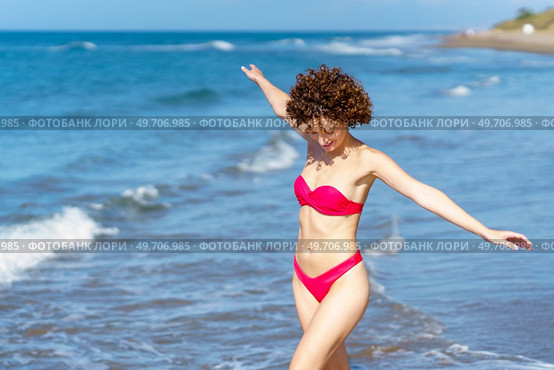 Delighted young fit female with dark curly hair, in red bikini smiling and looking down while walking in wavy sea with outstretched arms during summer holidays holidays. Joyful young lady in swimwear walking in sea with outstretched arms. Стоковое фото, агентство Ingram Publishing / Фотобанк Лори