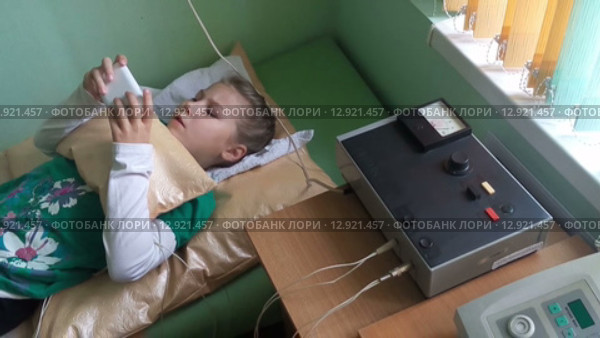 Девочка школьного возраста лежит на кушетке и играет с телефоном во время процедуры электрофореза в детской поликлинике (2015 год). Редакционное видео, видеограф Кекяляйнен Андрей / Фотобанк Лори