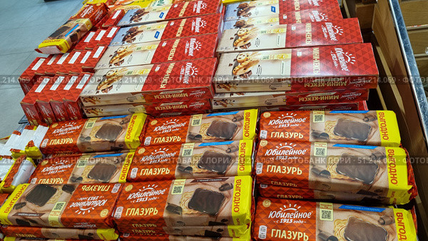 Display of confectionery products in a store. In the foreground are glazed Yubileiny cookies, packed in bright brown and yellow packages with the inscription "Bargain". On the shelves above are red packages of Yubileiny mini-muffins, as well as boxes of cookies and mini-muffins with chocolate chips. The packages are arranged in neat stacks, creating a rich and attractive composition for customers. The photo conveys the atmosphere of the supermarket, demonstrating the variety of sweet products and focusing attention on the Yubileiny brand. Редакционное фото, фотограф Анна Кривицкая / Фотобанк Лори