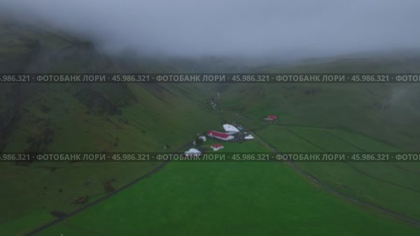 Дрон пролетает сквозь облака среди зелёных лугов и гор в Исландии, проходя над домами, расположенными рядом с водопадом, и запечатлевая величественный и умиротворённый пейзаж. Стоковое видео, видеограф Yana Veretennikova / Фотобанк Лори