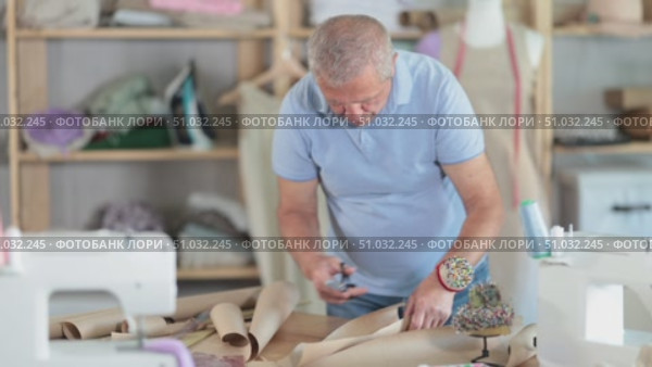 Elderly male tailor cutting out pattern from paper with scissors in sewing workshop. Стоковое видео, видеограф Яков Филимонов / Фотобанк Лори