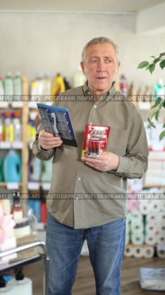 Elderly man is standing in a supermarket and holding razors from different manufacturers in hands, he is reading the instructions and manufacturing materials. Mature man chooses a trimmer and an electric shaver. Стоковое видео, видеограф Яков Филимонов / Фотобанк Лори