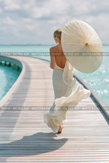 Elegant woman in flowing white dress walking on tropical boardwalk holding a parasol, surrounded by turquoise water under soft sunlight, viewed from behind. Стоковое AI-изображение, AI-художник Катерина Бондарец / Фотобанк Лори