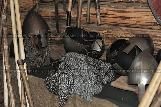 Elements of medieval military equipment, including metal helmets, chain mail and weapons. One of the helmets has a visor and a chain mail aventail, which is typical for medieval knights or warriors of Ancient Rus'. The other helmets have an open design and leather straps for fixation. In the foreground lies a chain mail shirt made of many metal rings. In the background, you can see various axes, clubs and other types of bladed weapons hanging on a wall made of logs. The whole scene resembles a military arsenal or a museum exhibition associated with historical reconstruction. Стоковое фото, фотограф Анна Кривицкая / Фотобанк Лори