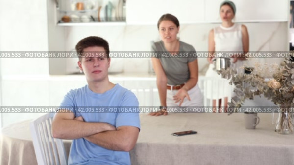 Emotionally tense distressed young guy sitting in home kitchen against blurred background of angry mother and sister standing in back and expressing dissatisfaction. Concept of family problems. Стоковое видео, видеограф Яков Филимонов / Фотобанк Лори