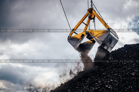 Excavator crane is actively unloading a coal bucket at a mining location where large piles of coal are visible. The heavy machinery, equipped with a dual bucket attachment, is positioned high above the coal mound. Стоковое фото, фотограф Владимир Разгуляев / Фотобанк Лори