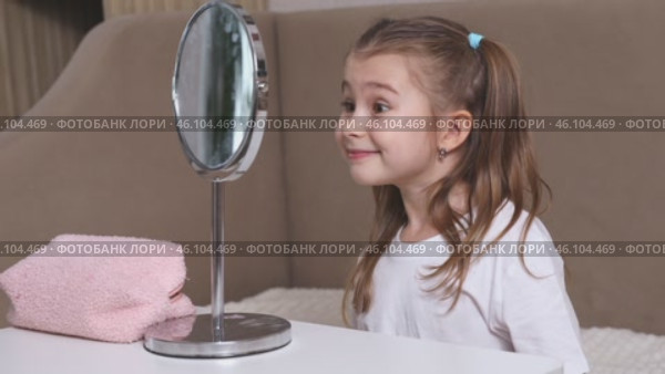 Fashion little girl in a white T-shirt is sitting at the dressing table in front of the mirror, smiling and winking her eyes. Advertising of children's cosmetics concept. Footage for kids blog. Стоковое видео, видеограф Ирина Михайличенко / Фотобанк Лори