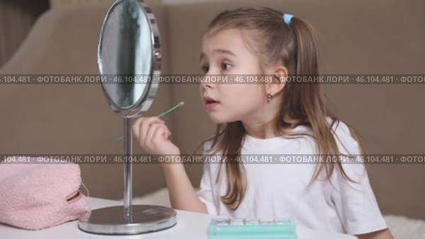 Fashion little girl in a white T-shirt is sitting at the dressing table in front of the mirror and applying makeup with eye shadow. Advertising of children's cosmetics concept. Footage for kids blog. Стоковое видео, видеограф Ирина Михайличенко / Фотобанк Лори