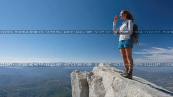 Female hiker standing on mountain peak, gesturing while absorbing expansive panoramic landscape with rolling hills, valleys stretching beneath luminous blue sky and distant horizon. Стоковое AI-видео, аниматор Юрий Поздников / Фотобанк Лори