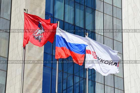 Флаг города Москвы, флаг Российской Федерации и флаг с логотипом компании "Лукойл" в ясную погоду на фоне фасада здания (Штаб-квартира российской нефтяной компании «Лукойл», Сретенский бульвар, 11). Город Москва (2018 год). Редакционное фото, фотограф Овчинникова Ирина / Фотобанк Лори