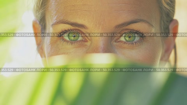 Focused close-up on female expressive green eyes peering through blurred green foliage, emphasizing her gaze and connection with nature. Стоковое AI-видео, аниматор Елена Никишина / Фотобанк Лори