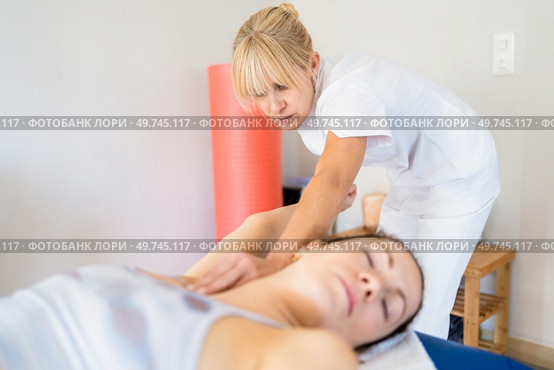 Focused female physiotherapist in white uniform massaging armpit of woman lying on couch during osteopathy session. Professional osteopath manipulating arm of patient. Стоковое фото, агентство Ingram Publishing / Фотобанк Лори