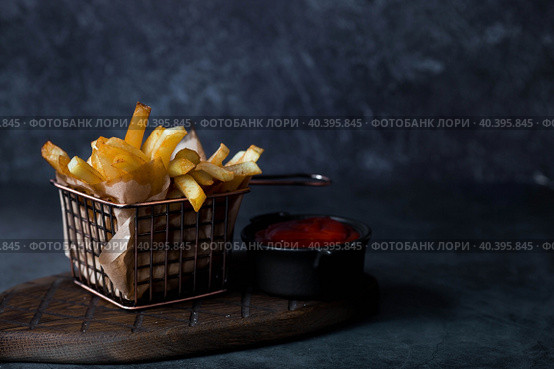 French fries in a metal basket with salt and ketchup on a stylish board on a dark background. Fried potatoes. The concept of fast food and junk food. Стоковое фото, фотограф Photojuli / Фотобанк Лори