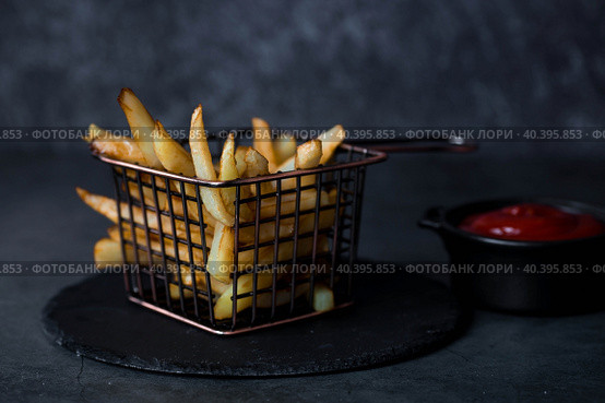 French fries in a metal basket with salt and ketchup on a stylish board on a dark background. Fried potatoes. The concept of fast food and junk food. Стоковое фото, фотограф Photojuli / Фотобанк Лори