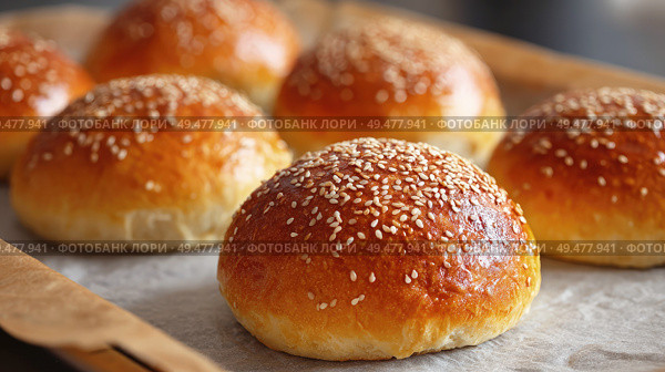 Freshly baked golden sesame seed buns on a parchment-lined tray, showcasing a glossy crust, soft texture, and appetizing homemade appearance in a close-up shot. Стоковое AI-изображение, AI-художник Катерина Бондарец / Фотобанк Лори