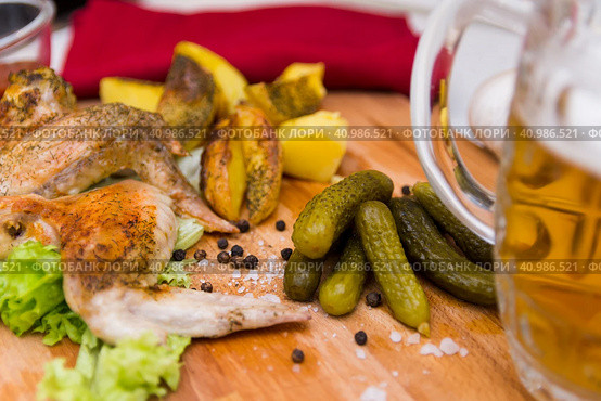 Fried chicken wings served on a wooden Board with Idaho potatoes, pickled gherkins and fresh lettuce leaves. A mug of beer is next to it. жареные куриные крылышки подаются на деревянной доске с картофелем Айдахо, маринованными корнишонами и свежими листьями салата. Рядом с ним стоит кружка пива. Стоковое фото, фотограф Дмитрий Березнев / Фотобанк Лори