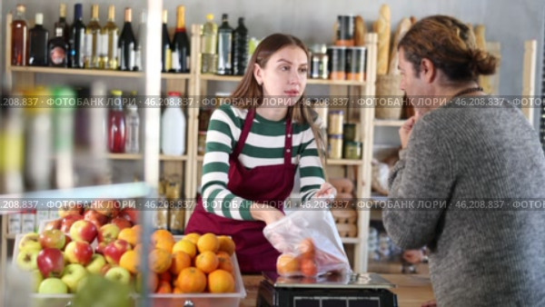 Friendly smiling young saleswoman in striped jumper and apron chatting warmly with male customer while weighing fresh tangerines in cozy grocery store. Стоковое видео, видеограф Яков Филимонов / Фотобанк Лори