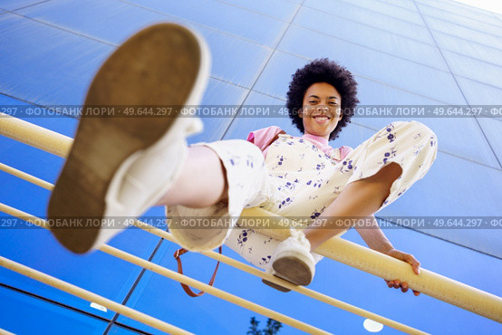 From below full body of positive African American female in overall looking at camera while sitting on fence near glass building. Smiling black woman sitting on railing. Стоковое фото, агентство Ingram Publishing / Фотобанк Лори