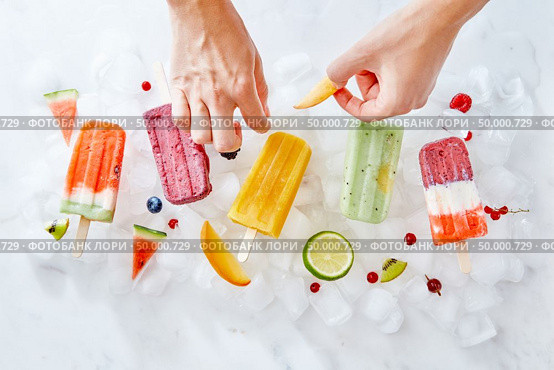 Fruit berry ice lolly on ice cubes. A woman’s hand decorates ice cream with pieces of fresh fruit fruit. Flat lay. The woman’s hands decorate the ice cream on a stick with bits of juicy fruit. Summer dessert. Flat lay. Стоковое фото, агентство Ingram Publishing / Фотобанк Лори