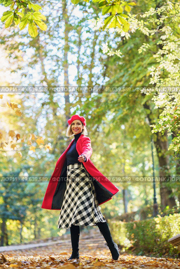 Full body of active female in red coat and beret looking at camera while standing on pathway on autumn day. Smiling woman in autumn park. Стоковое фото, агентство Ingram Publishing / Фотобанк Лори