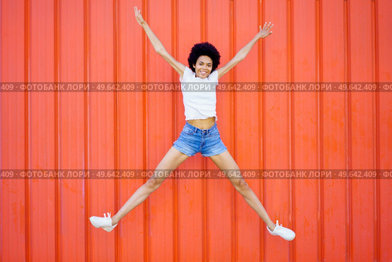 Full body of delighted African American female in casual wear looking at camera while jumping with raised arms against red background. Happy black woman jumping on street. Стоковое фото, агентство Ingram Publishing / Фотобанк Лори
