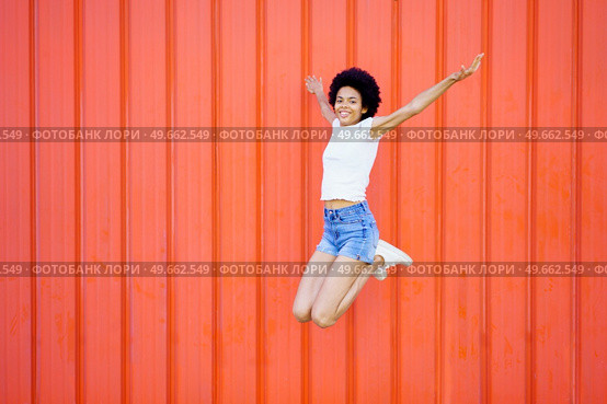 Full body of smiling African American female in casual outfit looking at camera while jumping with outstretched arms and bent knees against red background. Carefree black woman jumping near wall. Стоковое фото, агентство Ingram Publishing / Фотобанк Лори