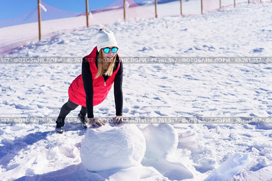 Full body of smiling young female blonde in beanie cap polarized sunglasses, looking away while leaning on hands and doing push ups on rock against wooden pillars fence in daylight. Happy woman warming up on snow terrain in winter. Стоковое фото, агентство Ingram Publishing / Фотобанк Лори