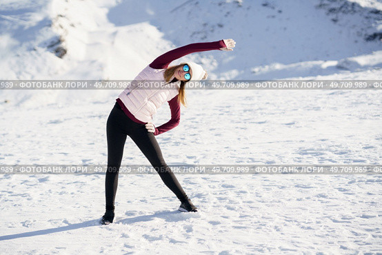 Full body of young female in sportswear and eyeglasses stretching arm overhead to one side while practicing yoga exercise on snowy slope in winter at daylight. Slim woman in sportswear stretching arm sideway in snowy mountains. Стоковое фото, агентство Ingram Publishing / Фотобанк Лори
