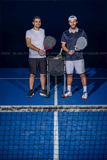 Full length portrait of two sporty young men in sportswear standing on indoor court with paddle rackets in hands after match. Стоковое фото, агентство Ingram Publishing / Фотобанк Лори
