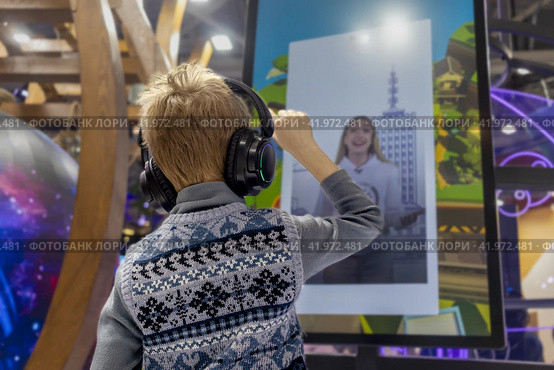 Generation Alpha boy wearing wireless headphones interacts with an interactive screen. Стоковое фото, фотограф Алла Чесноковва / Фотобанк Лори