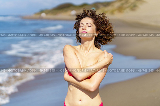 Gorgeous young female redhead relaxing with hands crossed, and touching bare shoulder while facing sun with eyes closed in daylight and standing in beach against blurred sea and sky. Young woman standing on sandy beach on sunny day. Стоковое фото, агентство Ingram Publishing / Фотобанк Лори