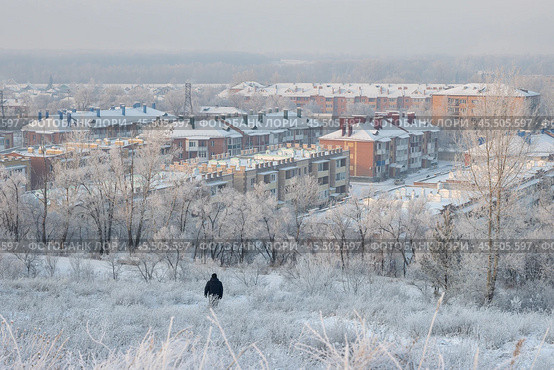 Город Абакан, Республика Хакасия, Сибирь, Россия. Зимний городской пейзаж. По заснеженному склону горы к городу спускается человек. Вид сверху на улицы и дома сибирского города. Снег на крышах зданий. Холодная погода. Морозный туман. На деревьях изморозь. Микрорайон Нижняя Согра. Стоковое фото, фотограф Андрей Степанов / Фотобанк Лори