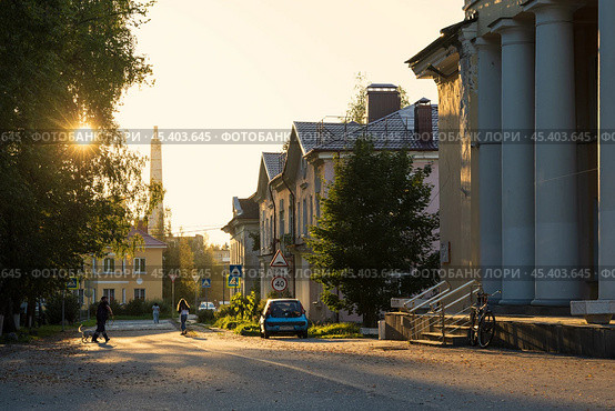 Городской посёлок Мга, Ленинградская область, Россия. Красивый вечерний городской пейзаж. Вид на улицу, освещённую солнцем. Солнечные лучи пробиваются через кроны деревьев. По асфальтовой дороге идут прохожие. Повседневная жизнь в регионах России. Редакционное фото, фотограф Андрей Степанов / Фотобанк Лори
