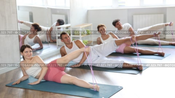 Group of young adults maintaining physical health practicing pilates with elastic band, lying on side and stretching legs at fitness center. Стоковое видео, видеограф Яков Филимонов / Фотобанк Лори
