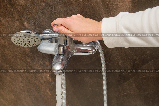 Hand adjusting shower faucet with running water in a bathroom. Close-up of shower control and showerhead against a dark brown wall. Household plumbing and water flow concept. Стоковое фото, фотограф Андрей Журавлев / Фотобанк Лори