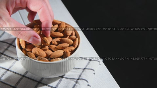Hand picking an almond from a white ceramic bowl overflowing with almonds, resting on a checkered tablecloth atop a light wooden table, highlighting healthy snacking options, copy space. Стоковое видео, видеограф Ольга Ильина / Фотобанк Лори