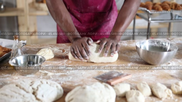 Hands of professional African baker in maroon apron kneading dough on floury wooden table, filled with baking tools and ingredients, in artisan bakery shop. Стоковое видео, видеограф Яков Филимонов / Фотобанк Лори