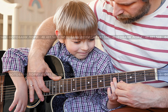 Handsome dad teaches his cute son to play the guitar sitting on sofa in the living room at home. Happy Father's Day. Father teaching his child to play guitar. Kid Learning music skill. Art education. Стоковое фото, фотограф Марина Демидюк / Фотобанк Лори