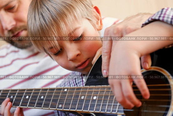 Handsome dad teaches his cute son to play the guitar sitting on sofa in the living room at home. Happy Father's Day. Father teaching his child to play guitar. Kid Learning music skill. Art education. Стоковое фото, фотограф Марина Демидюк / Фотобанк Лори