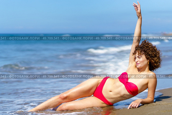 Happy curly woman in bikini with slim body raising arm while lying on beach of ocean. Slim woman in bikini suntanning on sea shore. Стоковое фото, агентство Ingram Publishing / Фотобанк Лори