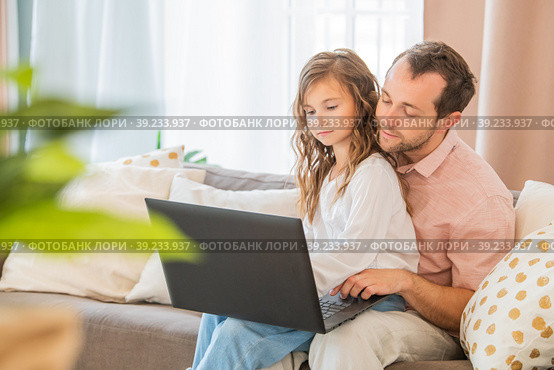 Happy family, daughter sitting on her father's lap on couch at home looking at laptop monitor together. Стоковое фото, фотограф Мария Мороз / Фотобанк Лори