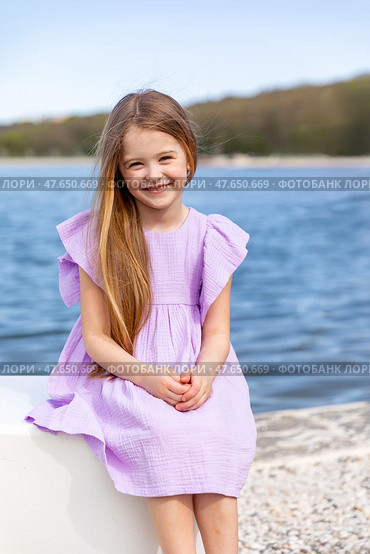 Happy little girl with long blonde hair, in lilac summer dress, sitting on the beach with blue water background. Children's Day. Vacation concept. Space for text. Advertising for tour operators. Стоковое фото, фотограф Ирина Михайличенко / Фотобанк Лори