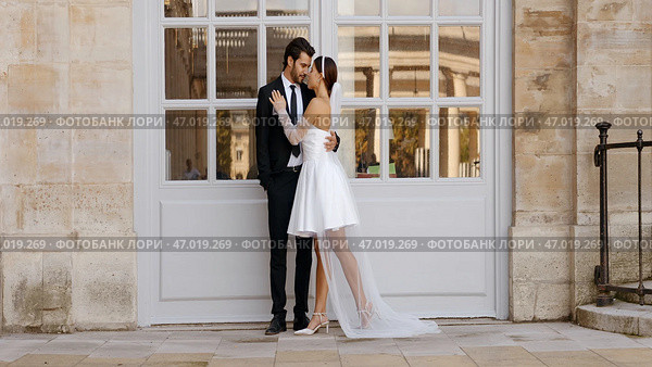 Happy newlyweds embracing each other by the white entrance door with windows. Стоковое фото, фотограф David Kashakhi / Фотобанк Лори