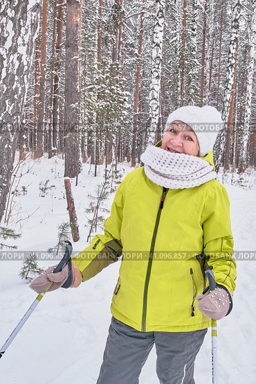 Happy smiling senior woman in yellow jacket, white knitted hat and scarf with ski poles over background of winter snowy forest. Стоковое фото, фотограф Ирина Кононова / Фотобанк Лори