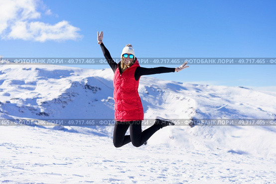 Happy young female in warm clothes cap polarized sunglasses looking at camera, while jumping in air with bent legs and stretched arms in daylight against snowy mountains and blue sky. Carefree young woman jumping with raised arms on snow terrain. Стоковое фото, агентство Ingram Publishing / Фотобанк Лори