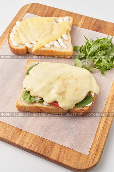 High angle of wooden board with parchment paper and toasted bread slices with cheese and vegetables and sauce before combining into delicious sandwich.. Preparation of tasty sandwich with cheese. Стоковое фото, агентство Ingram Publishing / Фотобанк Лори